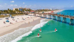 "Surfers dressed as Santa Claus riding waves near the decorated Cocoa Beach Pier, with holiday beachgoers, adorned palm trees, and lit boats in the background, under the warm winter sun in Cocoa Beach, Florida."