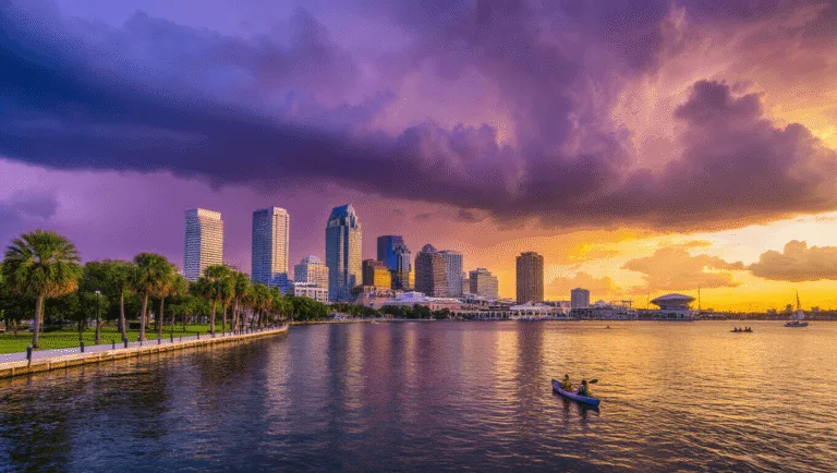 "Tampa cityscape at sunset with skyline reflected in Tampa Bay, palm trees, kayakers, sailboat, Tampa Museum of Art, Raymond James Stadium, lush parks along the shoreline, and storm clouds building in the distance"