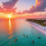 "Aerial sunset view of Anna Maria Island, with turquoise Gulf of Mexico and Tampa Bay waters meeting at Bean Point, kayakers and dolphins visible in water, beach umbrellas and beach houses along white sandy shore, framed by palm trees and a trolley on the coastal road."
