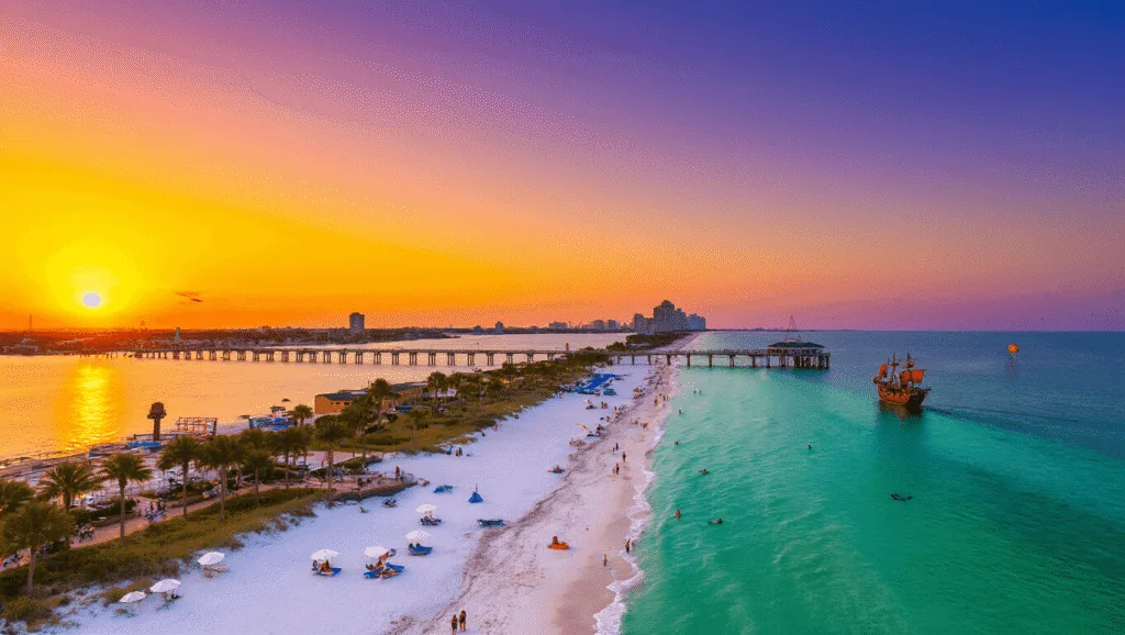 "Aerial view of Clearwater Beach at sunset with Pier 60, palm trees, white umbrellas, dolphins, parasailers, beachgoers, a pirate ship cruise, and Clearwater skyline"