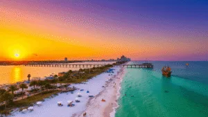 "Aerial view of Clearwater Beach at sunset with Pier 60, palm trees, white umbrellas, dolphins, parasailers, beachgoers, a pirate ship cruise, and Clearwater skyline"