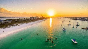 "Dramatic drone shot of Destin's coastline at sunset, showcasing emerald-green waters, pristine sandy beaches, Henderson Beach State Park's boardwalk, parasailers, dolphins, fishing boats, and HarborWalk Village with beachside activities and water sports."