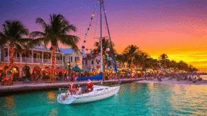 "Christmas decorations and palm trees on Duval Street in Key West during a vibrant December sunset with a lit sailboat in foreground and street performers in the background"