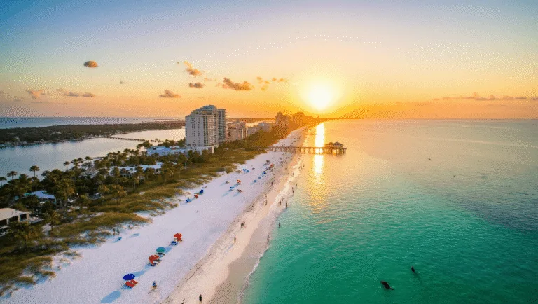 "Aerial view of Panama City Beach at sunset in May, featuring emerald waters and white sand, scattered beachgoers, colorful umbrellas, swaying palm trees, distant Pier Park, playful dolphins, and a parasailing boat, conveying serenity and spaciousness."