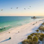 "Aerial view of Panama City Beach in February with sparse crowds, emerald-green waters, dolphins near the shoreline, beachgoers walking along the water's edge, iconic pier in the distance, birds overhead, St. Andrews State Park and modern beachfront architecture."