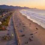 "Aerial view of Seaside, Oregon's coastline at sunset with families, surfers, and horseback riders, featuring the promenade, Lewis & Clark monument, and Captain Kid Amusement Park."