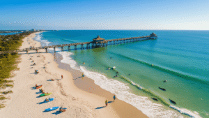 "Aerial view of Cocoa Beach in winter featuring golden sand, surfing waves, iconic pier, kayakers in Banana River Lagoon, dolphins, manatees, beachgoers, and stretching coastline of Space Coast."