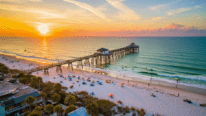 "Aerial view of Jacksonville Beach at sunset, featuring a fishing pier, surfers, colorful beach umbrellas, beachfront buildings, and palm trees"