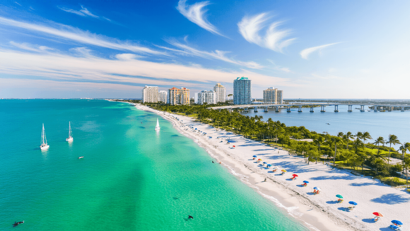 "Aerial view of Sarasota's coastline and Siesta Key beaches with turquoise waters, sailboats, palm trees, beach umbrellas, the John Ringling Causeway Bridge, and the Marie Selby Botanical Gardens under an April mid-afternoon sun."