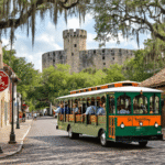 Why the St. Augustine Trolley Tour Is Your Secret Weapon for Conquering America's Oldest City "Old Town Trolley in bright green and orange rolling down cobblestone streets in St. Augustine, Florida with Spanish colonial buildings, Castillo de San Marcos in the background, tourists on board, and a trolley stop sign showing '22 stops' in the foreground"