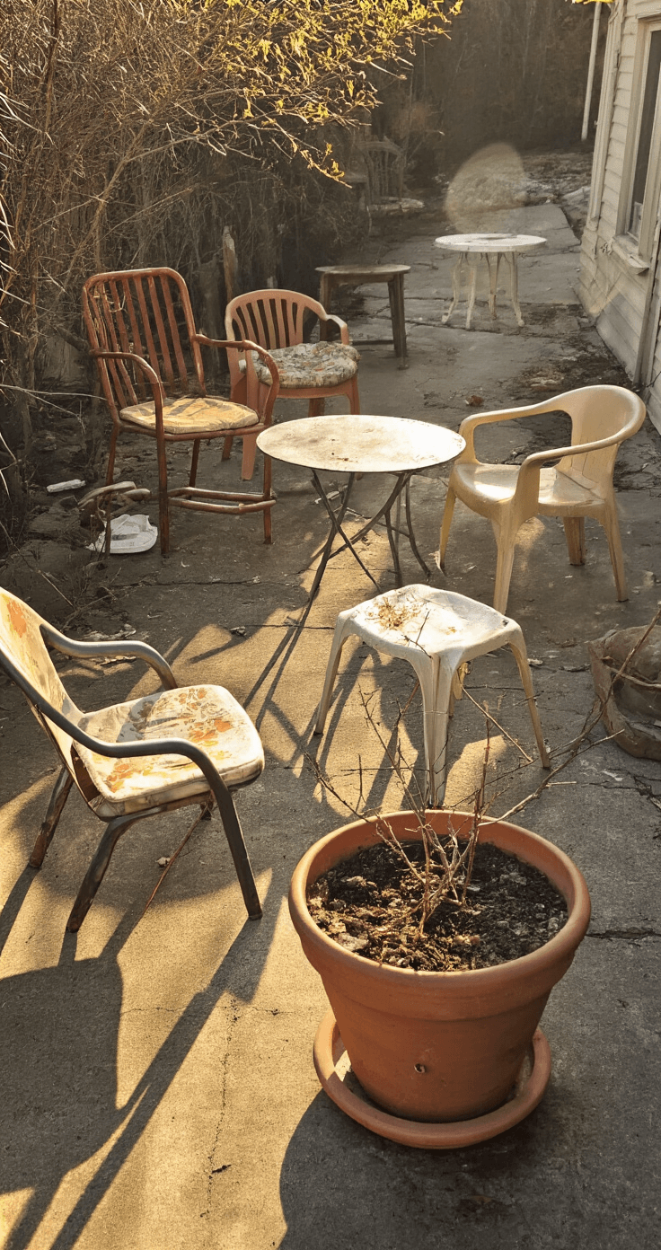 How I Finally Made My Outdoor Space Actually Usable This Spring (Without Breaking the Bank) A cluttered spring patio with mismatched outdoor furniture, including a rusty 80s metal chair, faded plastic chairs, and a wobbly bistro set, all on weathered concrete. A cracked terracotta pot holds a dead plant in the foreground, while peeling paint and mildew-stained cushions add to the neglected atmosphere, captured in harsh afternoon light with muted colors.