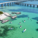 Why Fort De Soto Park Is Florida's Best-Kept Paddleboarding Secret (And How to Experience It Like a Pro) "Aerial view of Fort De Soto Park with paddleboarders in turquoise waters, white sand beaches, wooden docks, mangrove shores, Pinellas Bayway bridges, seagrass beds, dolphins, kayaks, pelicans, and an osprey nest."