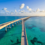The Seven Mile Bridge Drive That Changed Everything I Thought About Florida Road Trips "Panoramic view of Seven Mile Bridge in Florida, with old pedestrian walkway beside modern highway bridge, viewed over turquoise waters with visible ocean floor and distant dolphins, during golden hour"